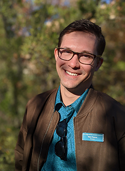 Nick smiles while standing in front of green foliage, in full sunlight